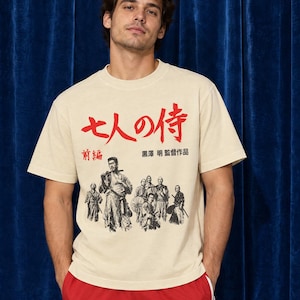 May include: A beige short-sleeved t-shirt featuring a black and white graphic of seven samurai figures with large red Japanese text above and smaller black text below. The shirt is paired with bright red athletic shorts with white side stripes.