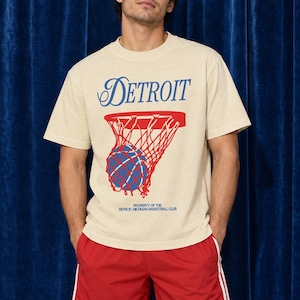 Könnte beinhalten: Ein beiges Kurzarm-T-Shirt mit dem blauen Schriftzug DETROIT über einer Grafik eines roten Basketballkorbs mit einem blauen Basketball sowie dem kleineren Text PROPERTY OF THE DETROIT, MICHIGAN BASKETBALL CLUB, kombiniert mit roten Sportshorts.