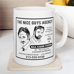 May include: White ceramic mug with the text "THE NICE GUYS AGENCY" and illustrations of two men. The mug also includes the text "ALL CASE TYPES" and contact information. The mug is on a beige coaster.