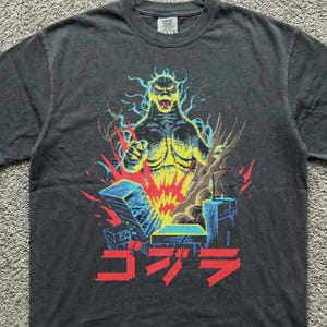 Op de afbeelding: Donkergrijs t-shirt met een Godzilla-afbeelding. Het ontwerp bevat een geel en blauw monster, rode vlammen en Japanse tekst. Het shirt heeft een vintage, verweerde uitstraling.