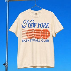 May include: Cream-coloured t-shirt featuring the text "New York Basketball Club" in blue script and block letters. Below the text are three orange basketballs and horizontal red lines. The shirt is hanging on a metal rack.