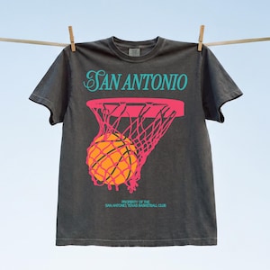 Puede incluir: Camiseta gris oscuro con un diseño de baloncesto. La camiseta presenta el texto "SAN ANTONIO" en escritura turquesa sobre una red de baloncesto rosa con un balón. Debajo, el texto "PROPERTY OF THE SAN ANTONIO, TEXAS BASKETBALL CLUB."