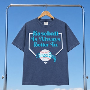 Könnte beinhalten: Marineblaues T-Shirt mit dem Text "Baseball Is Always Better In Tampa Bay" in Weiß und Türkis. Das Design beinhaltet einen Baseball und eine Homeplate-Umrandung. Das Shirt hängt an einem Holzbügel.