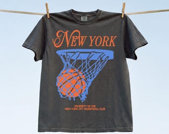 May include: A charcoal gray short-sleeve t-shirt featuring the orange text New York above a blue basketball hoop graphic with an orange basketball. Below the graphic is the orange text Property of the New York City Basketball Club.
