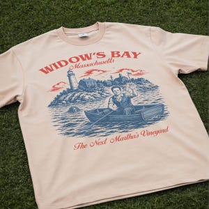 May include: A light beige t-shirt featuring a graphic print with the text "WIDOW'S BAY Massachusetts" and "The Next Martha's Vineyard". The illustration depicts a person rowing a boat near a lighthouse and buildings, with a creature in the water.