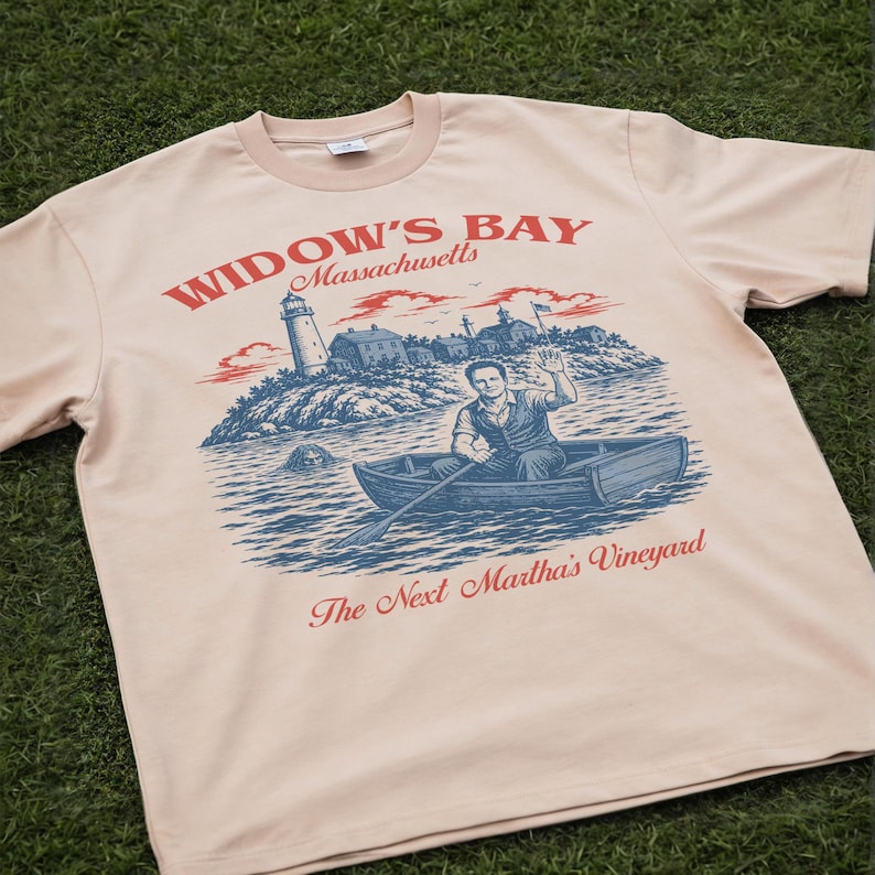 May include: A light beige t-shirt featuring a graphic print with the text "WIDOW'S BAY Massachusetts" and "The Next Martha's Vineyard". The illustration depicts a person rowing a boat near a lighthouse and buildings, with a creature in the water.