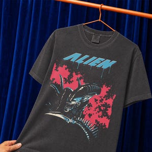 May include: A charcoal grey short-sleeve t-shirt featuring a graphic print of the Alien creature. The word ALIEN is printed in light blue above the creature, which is set against a rectangular pink background with a dark, textured pattern.