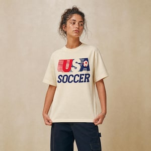 May include: A cream-colored short-sleeve t-shirt featuring a graphic print on the chest. The design includes the text USA in red and blue with horizontal lines, a small soccer ball icon, and the word SOCCER in bold blue capital letters below.