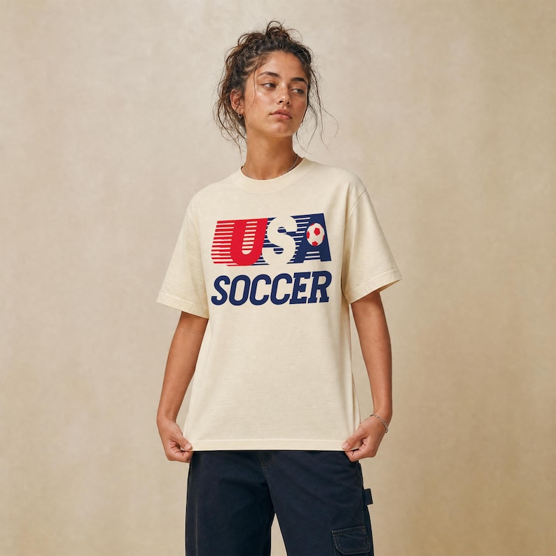 May include: A cream-colored short-sleeve t-shirt featuring a graphic print on the chest. The design includes the text USA in red and blue with horizontal lines, a small soccer ball icon, and the word SOCCER in bold blue capital letters below.
