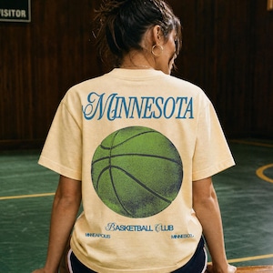 May include: A light yellow t-shirt with the word "MINNESOTA" in blue script above a green basketball graphic. Below the basketball, the text reads "BASKETBALL CLUB" and "MINNEAPOLIS".