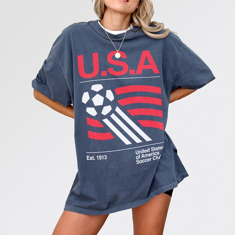 May include: A dark blue t-shirt with the text "U.S.A" in red, a soccer ball graphic, and red and white stripes. The shirt also includes the text "Est. 1913" and "United States of America Soccer Club".