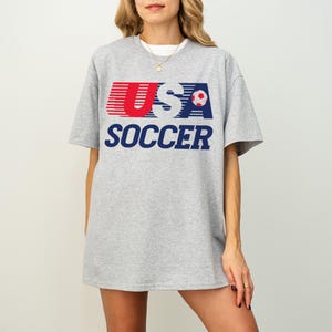 May include: A heather gray short-sleeve t-shirt featuring the text USA SOCCER in red, white, and blue. The design includes a small red and white soccer ball graphic integrated into the letter A. The shirt has a crew neckline and a relaxed fit.