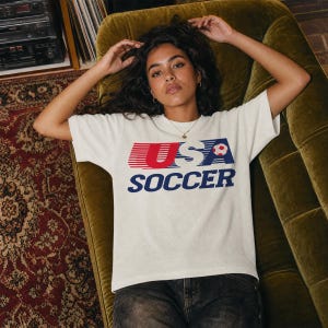 May include: A white short-sleeved t-shirt featuring a graphic print on the chest with the text USA in red and blue, the word SOCCER in blue, and a small soccer ball icon. The design includes horizontal red and blue motion lines.