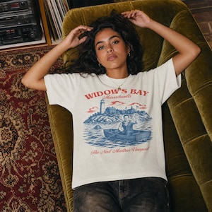 May include: An off-white short-sleeve t-shirt featuring a graphic print of a lighthouse on an island and a person in a rowboat. The text reads Widow's Bay Massachusetts and The Next Martha's Vineyard in red lettering with blue water and landscape.