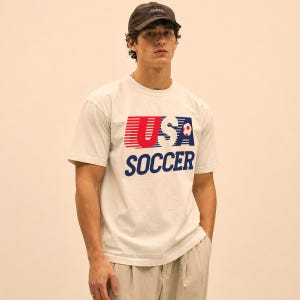 May include: A cream-colored short-sleeve t-shirt featuring a graphic print that reads USA SOCCER in red and blue with horizontal lines and a small soccer ball icon. The shirt is paired with a dark brown baseball cap with the word TOKYO embroidered.