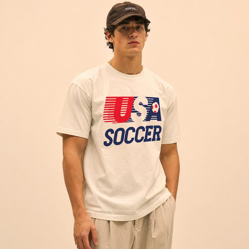 May include: A cream-colored short-sleeve t-shirt featuring a graphic print that reads USA SOCCER in red and blue with horizontal lines and a small soccer ball icon. The shirt is paired with a dark brown baseball cap with the word TOKYO embroidered.