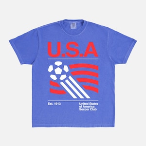 K&ouml;nnte beinhalten: Blaues T-Shirt mit einer Grafik mit den roten Buchstaben "U.S.A" &uuml;ber einem Fu&szlig;ball und rot-wei&szlig;en Streifen. Der Text "Est. 1913" und "United States of America Soccer Club" stehen darunter.