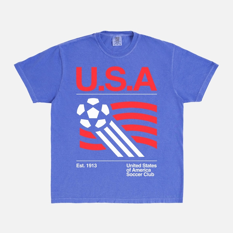 K&ouml;nnte beinhalten: Blaues T-Shirt mit einer Grafik mit den roten Buchstaben "U.S.A" &uuml;ber einem Fu&szlig;ball und rot-wei&szlig;en Streifen. Der Text "Est. 1913" und "United States of America Soccer Club" stehen darunter.