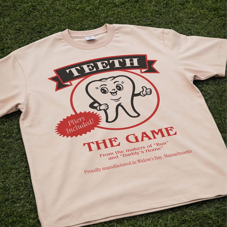 May include: A light beige t-shirt featuring a graphic of a cartoon tooth character inside a red circle. The text reads TEETH THE GAME, Pliers Included!, From the makers of Run and Daddy's Home, and Proudly manufactured in Widow's Bay, Massachusetts.