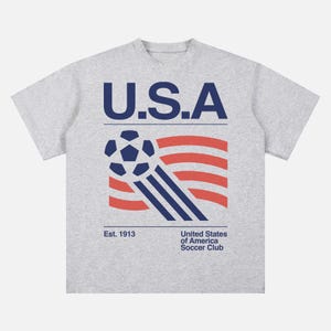 K&ouml;nnte beinhalten: Graues T-Shirt mit einem Grafikdesign, das einen Fu&szlig;ball, rot-wei&szlig;e Streifen und den Text "U.S.A." in Marineblau zeigt. Darunter stehen die Worte "Est. 1913" und "United States of America Soccer Club".