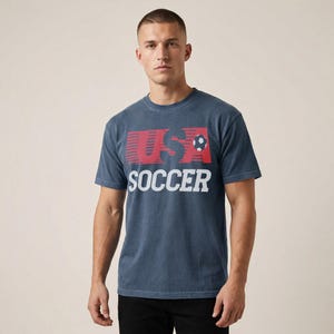 May include: A slate blue short-sleeve crew neck t-shirt featuring a graphic on the chest. The graphic displays the red text USA with horizontal motion lines and a small soccer ball icon, followed by the word SOCCER in large white block letters.