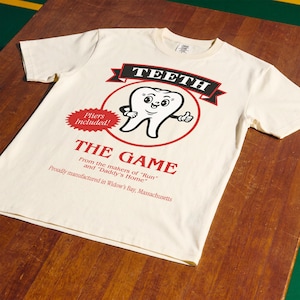 May include: A cream-colored short-sleeve t-shirt featuring a graphic of a cartoon tooth with arms and a face. The text reads TEETH, THE GAME, Pliers Included!, From the makers of Run and Daddy's Home, and Proudly manufactured in Widow's Bay, Massachusetts.