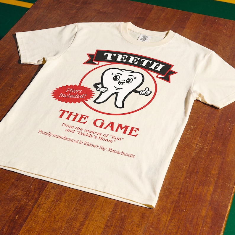 May include: A cream-colored short-sleeve t-shirt featuring a graphic of a cartoon tooth with arms and a face. The text reads TEETH, THE GAME, Pliers Included!, From the makers of Run and Daddy's Home, and Proudly manufactured in Widow's Bay, Massachusetts.