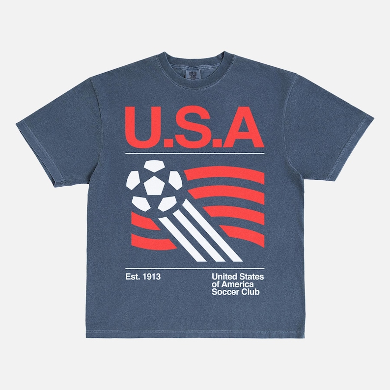 K&ouml;nnte beinhalten: Marineblaues T-Shirt mit einer Grafik eines Fu&szlig;balls und rot-wei&szlig;en Streifen, sowie dem Text "U.S.A." in Rot. Darunter steht "Est. 1913" und "United States of America Soccer Club."