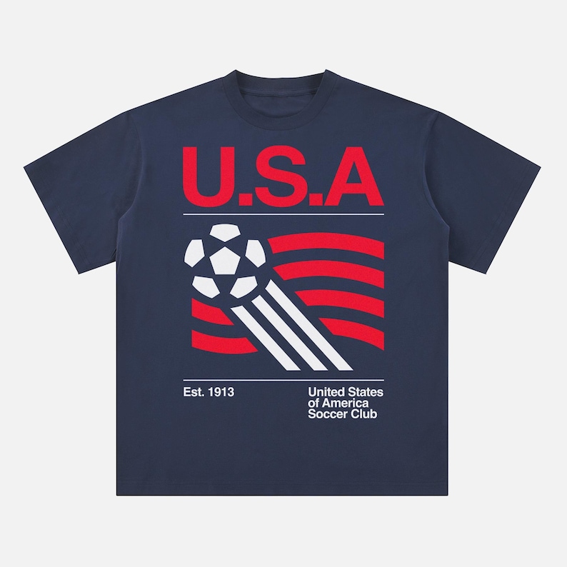 K&ouml;nnte beinhalten: Marineblaues T-Shirt mit rotem "U.S.A."-Schriftzug und einem Fu&szlig;ball-Design. Das Design umfasst rote und wei&szlig;e Streifen sowie den Text "Est. 1913" und "United States of America Soccer Club".