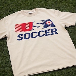 May include: A beige short-sleeved t-shirt featuring a graphic print on the chest. The design displays the text USA in red and blue with horizontal motion lines, a small red and white soccer ball icon, and the word SOCCER in bold blue letters.