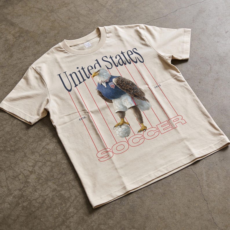 Op de afbeelding: Cr&egrave;mekleurig T-shirt met een afbeelding van een adelaar in een voetbaltenue, balancerend met een voetbal. De tekst "United States Soccer" is in marineblauw en rood op het shirt gedrukt.