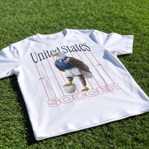 Op de afbeelding: Wit T-shirt met een gestileerde adelaar in voetbaltenue, die een voetbal vasthoudt. Het shirt heeft de woorden "United States" en "SOCCER" in marineblauw en rood, met rode strepen. Op een groene grasachtergrond.