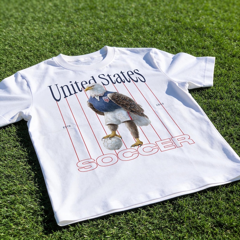 Op de afbeelding: Wit T-shirt met een gestileerde adelaar in voetbaltenue, die een voetbal vasthoudt. Het shirt heeft de woorden "United States" en "SOCCER" in marineblauw en rood, met rode strepen. Op een groene grasachtergrond.