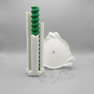 Op de afbeelding: Een 3D-geprinte assemblage met een verticale witte behuizing met een groene schroefdraadcomponent erin, vergezeld van een aparte witte ronde schijf met een centraal gat en een doorzichtige zeshoekige basisplaat met meerdere uitsparingen.
