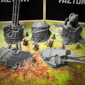 May include: A collection of gray miniature war game terrain pieces, including turrets, bunkers, and cannons. Several painted miniature figures are positioned around the terrain on a green and brown patterned surface. The pieces are likely for tabletop gaming.