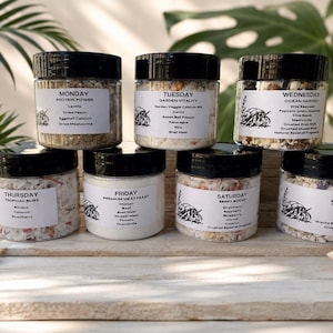 May include: Seven clear plastic jars with black lids, each containing a different dry food mix for hermit crabs. Each jar features a white label printed with a day of the week, a unique blend name, a list of ingredients, and a small hermit crab icon.