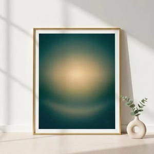 Peut inclure: Une impression d'art abstrait encadrée avec un cadre doré. L'œuvre présente un motif circulaire flou avec un dégradé de couleurs, passant d'un centre doré à des bords bleu canard foncé. Un petit vase avec de la verdure est à proximité.