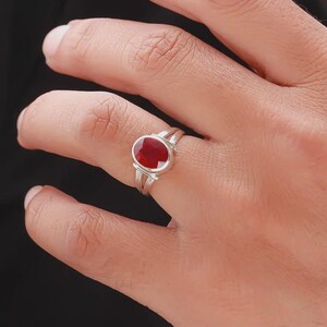May include: A silver ring featuring a large, oval-cut, deep red gemstone. The ring has a split shank design, with two parallel bands on each side of the gemstone. The gemstone is set in a bezel setting.