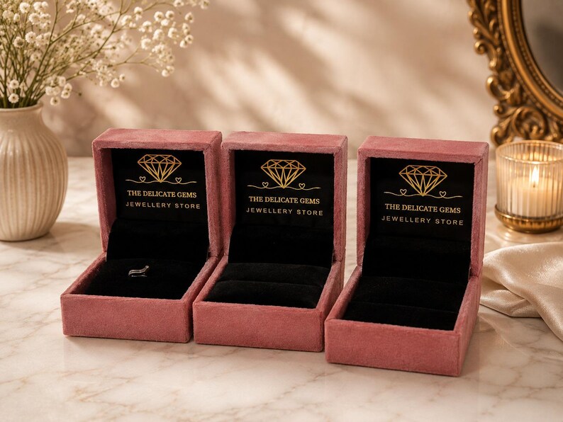 May include: Three dusty rose velvet jewelry boxes with black interiors. Each box features a gold diamond icon and the text THE DELICATE GEMS JEWELLERY STORE printed on the inside lid. One box contains a small silver ring on a black velvet insert.