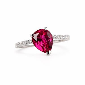 Può includere: Un anello color argento con una grande gemma rosso scuro a forma di pera come elemento centrale. La fascia è realizzata in materiale metallico ed è decorata con una serie di piccole pietre chiare e scintillanti su entrambi i lati.