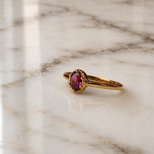 May include: A gold ring featuring a single oval-cut deep pink gemstone set in a scalloped bezel setting. The band is thin and polished, showcasing a smooth, reflective metallic finish that complements the vibrant color of the central stone.