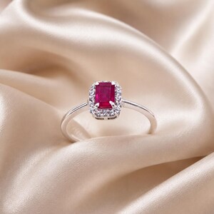 Può includere: Un anello in argento con una pietra centrale di rubino rosso rettangolare a taglio smeraldo, circondata da un alone di piccoli diamanti bianchi rotondi e trasparenti. La fascia è sottile e lucida, con un design classico e una gemma rossa.