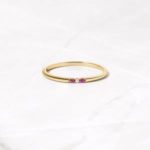 Peut inclure: Une fine bague en or avec trois petites pierres rondes serties en ligne horizontale. La pierre centrale est transparente et incolore, entourée de chaque côté par une pierre rose vif. Le métal présente une finition en or jaune poli.