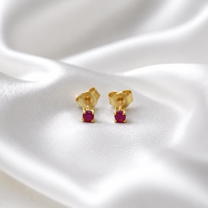 May include: A pair of stud earrings featuring small, round, vibrant pinkish-red gemstones set in polished yellow gold prongs. The earrings include matching yellow gold butterfly-style friction backs, showcasing a simple and classic design.
