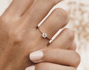 May include: A delicate gold ring with a single, round, clear gemstone. The ring is thin and sits on a finger, showcasing the minimalist design. The gemstone sparkles, catching the light. The ring is a piece of jewelry.