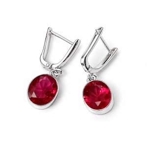 May include: A pair of silver-toned drop earrings featuring oval-shaped, faceted deep red stones set in smooth metal bezels. The earrings are attached to hinged lever-back closures, providing a secure fit for the dangling, polished gemstone pendants.
