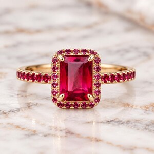 Ruby halo statement ring with an emerald cut ruby center stone and 925 sterling silver gold plated band photographed on a luxury white marble surface.