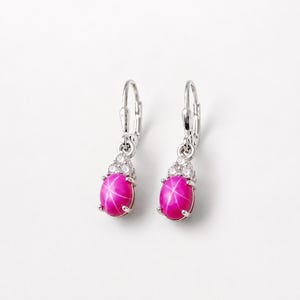 Puede incluir: Un par de pendientes colgantes con piedras rosas vibrantes en forma de estrella ovalada engastadas en metal plateado. Cada pendiente está decorado con tres pequeñas piedras claras brillantes en la parte superior y un cierre de palanca.