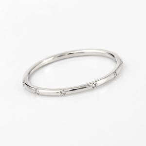 May include: A thin, polished silver-tone metal band ring featuring a series of small, round, clear stones set at regular intervals along the outer circumference of the band. The ring has a smooth, reflective finish and a simple, circular design.