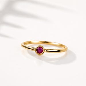 Può includere: Un sottile anello in oro con un singolo rubino rotondo incastonato a lunetta al centro. La fascia è liscia e lucida, caratterizzata da un design essenziale con una pietra rossa vivace tenuta saldamente all'interno della montatura circolare.